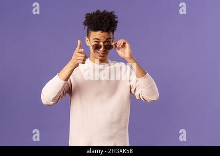 Portrait of cheeky handsome young man wearing sunglasses, take them off and show thumb-up, like or approval, recommendation sign Stock Photo