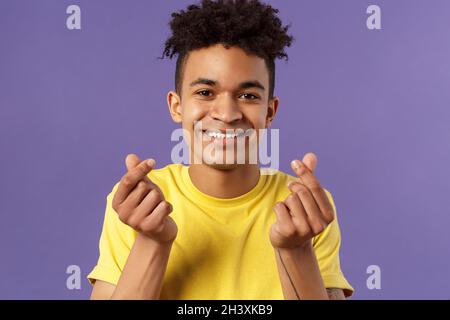 Close-up portrait of lovely cute young man with afro haircut, showing korean heart finger signs and smiling, express his love an Stock Photo