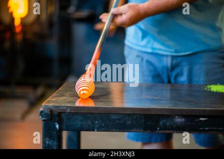 a worker is in the process of creating a glass vase and is rolling glass and adding decoration during the process of glass blowing Stock Photo