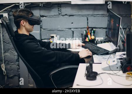 Young man programmer gamer sitting on chair with computer desktop near virtual reality goggles, vr glasses headset Stock Photo
