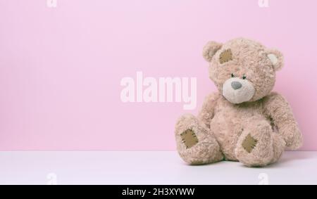 Brown teddy bear with patches sits on a white table, pink background Stock Photo