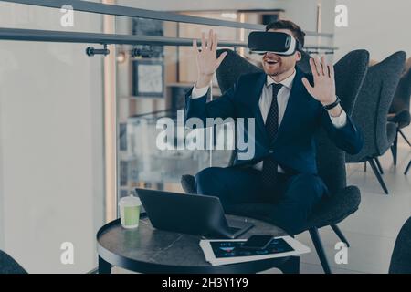 Businessman dressed in formal suit wearing VR goggles while working on laptop in cafe Stock Photo
