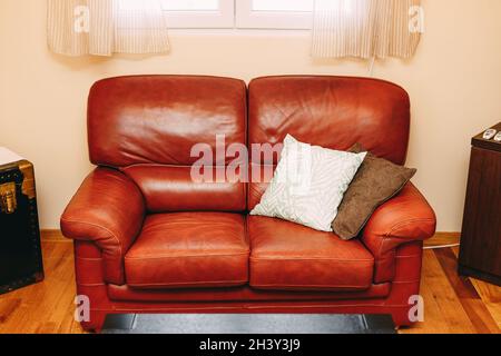 Folding sofa made of burgundy genuine leather with white and brown pillows under the window with beige curtains. Stock Photo