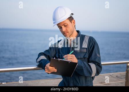 Focused young male port engineer in protective helmet and workwear taking notes on clipboard while standing near railing against sea Stock Photo