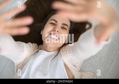 Photo from above of happy young positive woman lying on floor raising both hands up Stock Photo