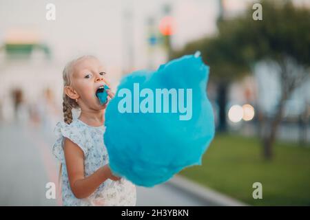 Little blond girl eating cotton candy and shows blue tongue in the park. Stock Photo