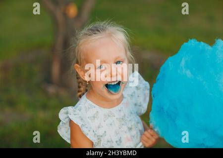 Little blond girl eating cotton candy and shows blue tongue in the park. Stock Photo