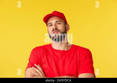 Close-up of confident handsome bearded delivery guy, male courier in red uniform cap and t-shirt, look tough and serious, standi Stock Photo