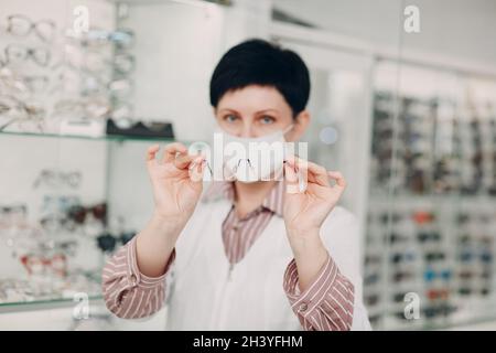 Happy Optician in Medical Face Mask Choosing Eyeglasses, female optometrist with glasses Stock Photo