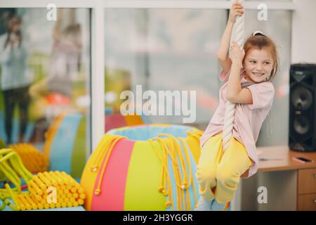 Kid girl doing exercises climbing tightrope in gym at kindergarten or elementary school. Children sport and fitness concept. Stock Photo