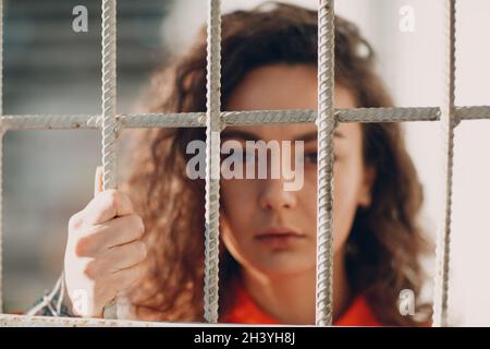 Young brunette curly woman in orange suit behind jail bars. Female in colorful overalls portrait. Stock Photo