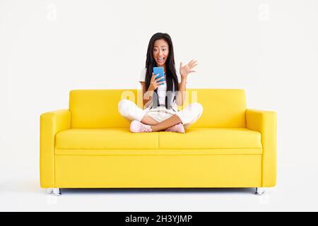 Emotional surprised asian female receiving good message on cell phone sitting on yellow couch Stock Photo