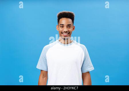 Charming friendly and polite dark-skinned guy with beard and afro hairstyle in trendy t-shirt smiling broadly gazing at camera f Stock Photo