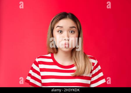 Close-up silly cute asian blond girl pouting making playful face, fold lips hold breath stand red background speechless, stay si Stock Photo