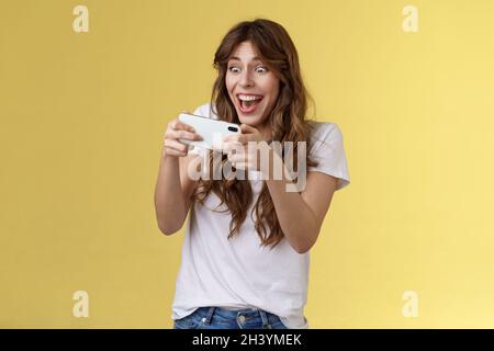 Happy upbeat playful enthusiastic curly-haired girl tempting playing stunning awesome smartphone game hold mobile phone horizont Stock Photo