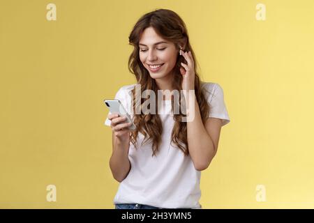 Tender feminine curly-haired female white t-shirt touch wireless earbud put earphone ear smiling pleased look smartphone screen Stock Photo