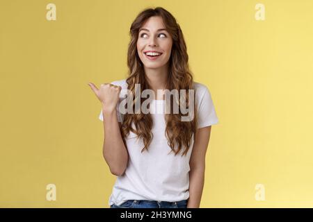 Something interesting right there. Curious lively enthusiastic charming woman glancing pointing thumb left smiling broadly discu Stock Photo