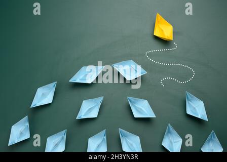 Group of blue paper boats are sailing in one direction, one yellow is sailing in the opposite direction. Stock Photo