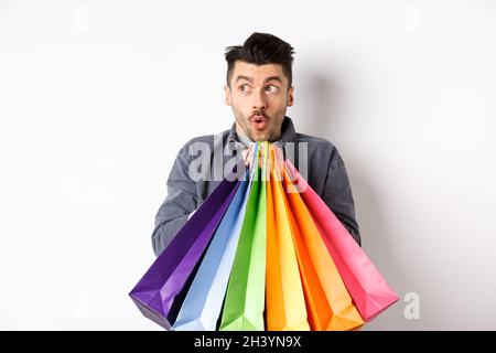 Excited man holding colorful shopping bags and looking left at special promo offer in store, standing on white background Stock Photo
