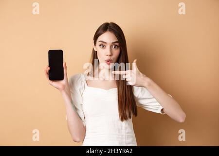 Excited woman showing news on screen, pointing at empty phone and looking surprised, standing on beige background Stock Photo