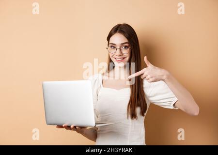 Smiling woman in glasses pointing finger at laptop screen, showing online promo, standing on beige background Stock Photo