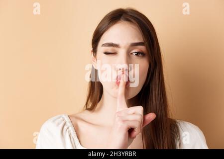 Close up of attractive woman shushing at camera, hiding secret, making hush sign and winking, show taboo sign, standing on beige Stock Photo