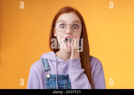 Omg no way. Attractive shocked wondered redhead amused hipster woman modern teenager drop jaw gasping wide eyes surprised standi Stock Photo