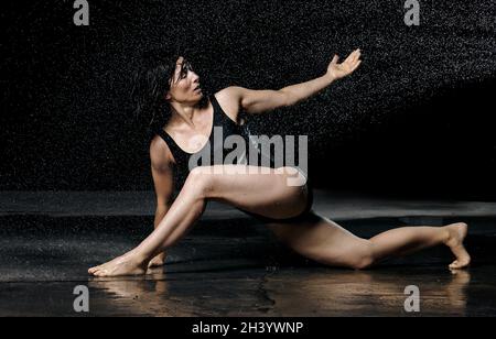 Woman with black hair lies on the ground under raindrops on a black background. Woman dressed in black bodysuit Stock Photo