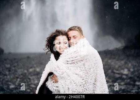 Wedding couple near Skogafoss waterfall. The bride and groom covered with a woolen blanket, where hugging. Snow is falling, fall Stock Photo