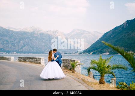 The bride and groom walk along the road near Perast and kiss, back view Stock Photo