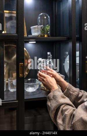 A woman holds a glass cup on the background of a cabinet with utensils. Stock Photo