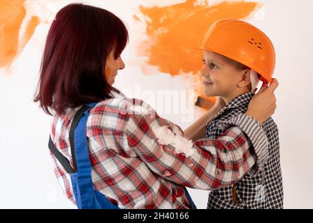Repair. Happy family mom and son in helmet painting home interior wall. Paint the wall orange. Portrait of a woman with a child, repairman. Stock Photo