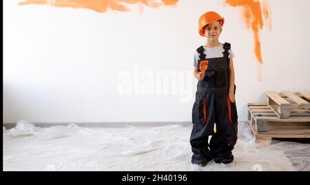 Caucasian boy in overalls and a protective helmet painting the wall with a brush in orange. Children are doing renovations in their new home. Stock Photo