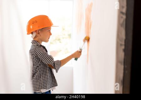 Caucasian child boy paints the wall with paint roller indoors doing repairs himself. children and home or apartment renovation. Kid in orange helmet Stock Photo