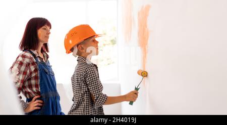 mom and son are painting the wall with orange paint. home renovation concept. A boy in an orange helmet paint a wall in a room or apartment a roller. Stock Photo