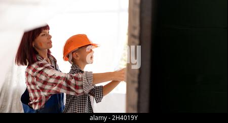 Mom teaches her son to do home repairs. Boy in orange helmet paint the wall in the room with a cushion helps the mother. A family makes renovations Stock Photo