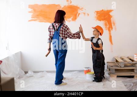 Mom and her little son are doing repairs in the room, they painted the wall orange with a brush and a roller. A family is delighted with the new room Stock Photo