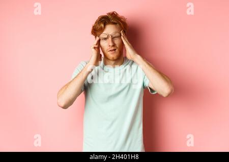Portrait of redhead man in crooked glasses touching head and feeling dizzy or nauseous, having hangover or headache, standing ov Stock Photo