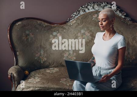 Millenial young woman with short blonde hair portrait sitting with laptop on vintage sofa. Stock Photo