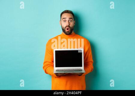 Handsome bearded man in orange sweater showing laptop screen, demonstrating promo, standing over light blue background Stock Photo