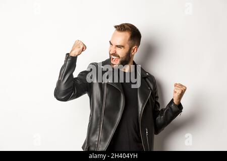 Confident bearded man celebrating victory, winning prize, making fist pump and rejoicing, wearing black leather jacket, white ba Stock Photo