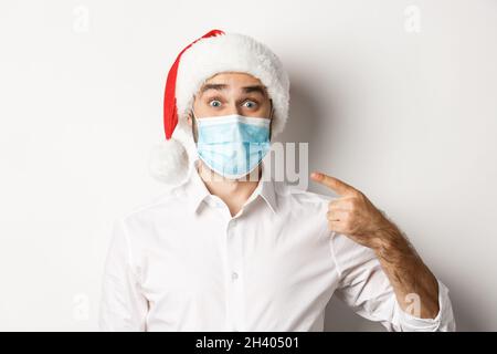 Concept of coronavirus, quarantine and winter holidays. Excited guy in santa hat pointing at his face mask, standing over white Stock Photo