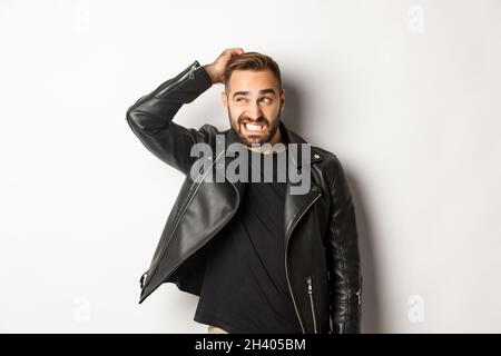 Confused macho guy in black leather jacket looking awkward and unsure, scratching head puzzled and stare left, white background Stock Photo