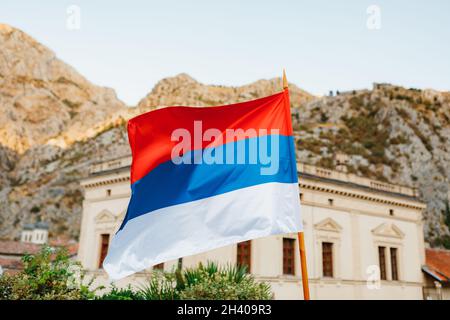 The flag of Serbia flies against the backdrop of a building and mountains on a sunny day. Stock Photo