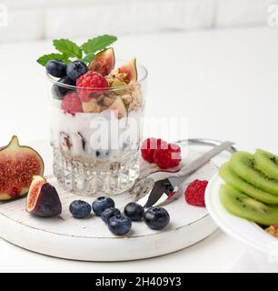 Transparent glass with granola poured with yogurt, on top of ripe raspberries, blueberries and figs on a white table. Healthy br Stock Photo