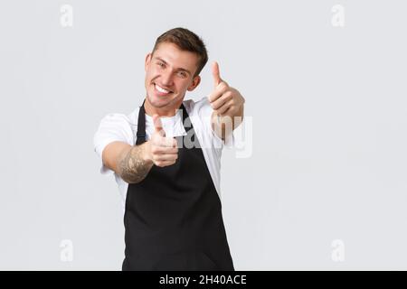 Employees, grocery stores and coffee shop concept. Cheerful handsome barista in cafe, store employee in black apron showing thum Stock Photo