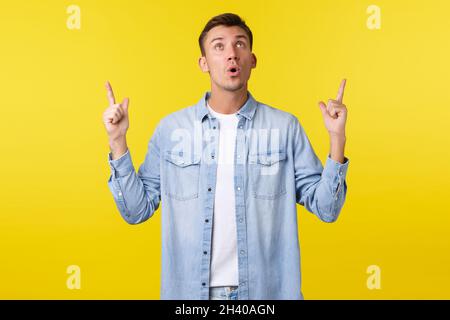 Surprised and excited handsome caucasian man saying wow, looking and pointing fingers up at awesome promo banner with big news, Stock Photo