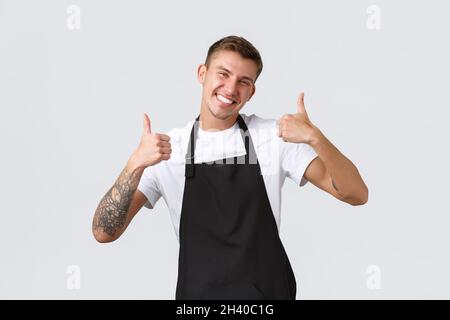 Employees, grocery stores and coffee shop concept. Handsome friendly barista in black apron, showing thumbs-up, inviting for cap Stock Photo