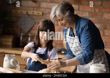 Cheerful granddaughter kid helping happy grandma to knead dough Stock Photo