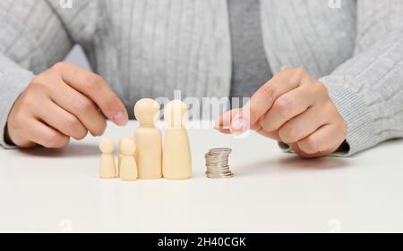 Female hand puts coins in a pile and wooden figurines of a family on a white table. Economy and investment concept, budget plann Stock Photo